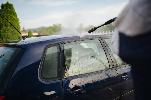 A person is washing a blue car in a residential driveway. Water spray from a pressure washer creates a mist under the warm sun, highlighting a clean and vibrant setting. How to Start a Pressure Washing Business in Texas with No Money